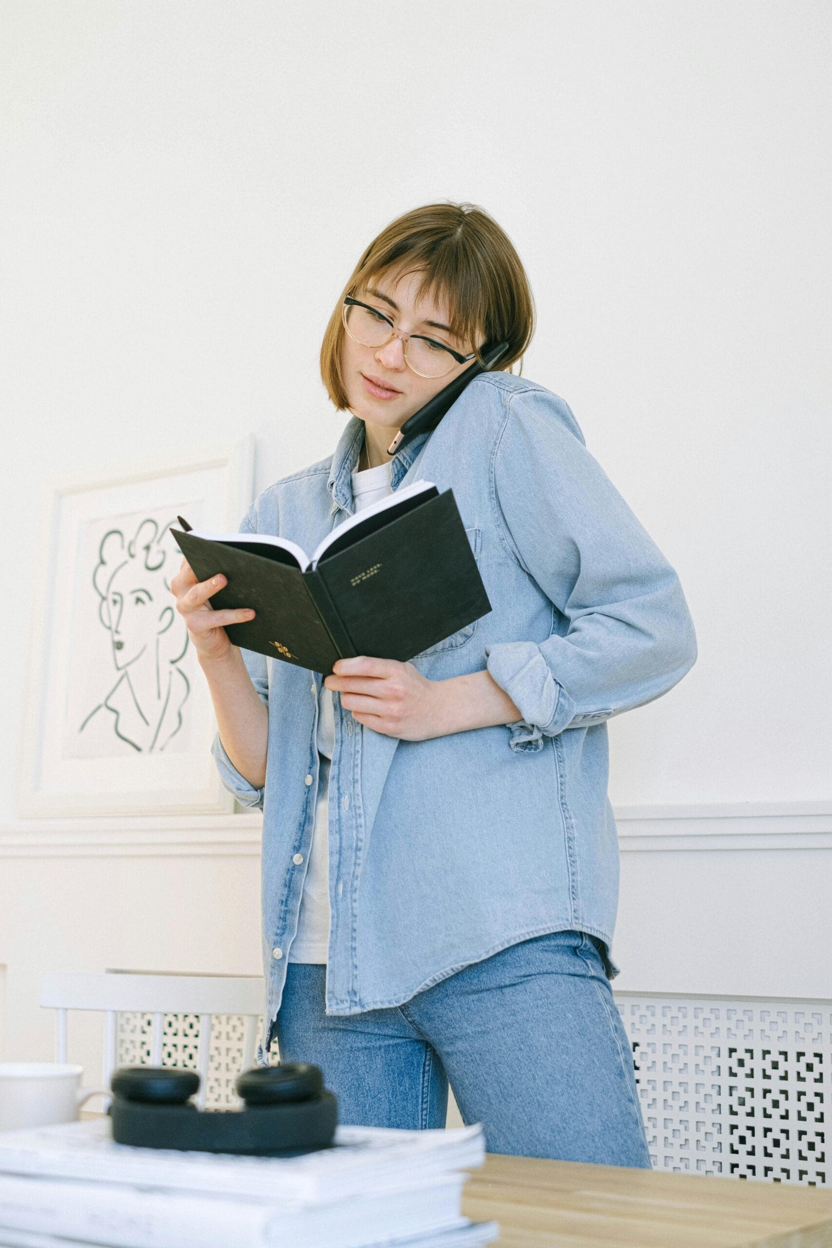 Young woman multitasking in a home office, holding an agenda and making a phone call.