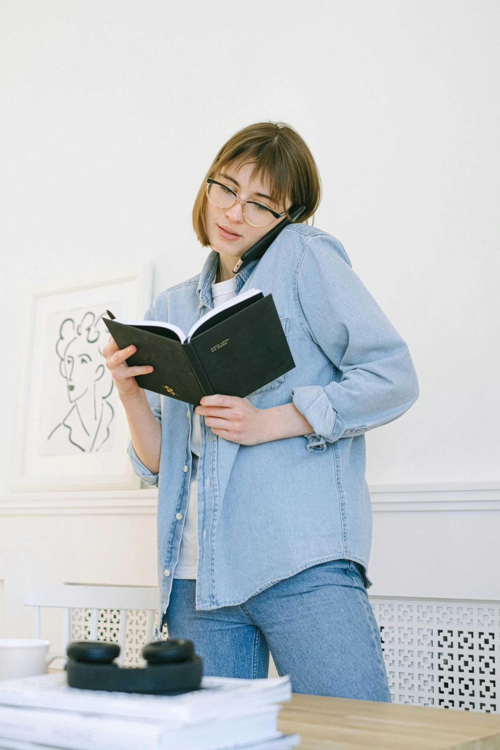 Young woman multitasking in a home office, holding an agenda and making a phone call.